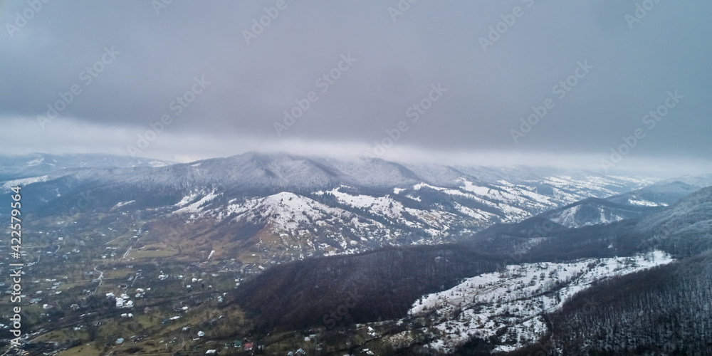 A beautiful landscape shot from a drone. A small village located near mountains and a forest covered with snow in foggy cloudy weather. Recreation, tourism concept.