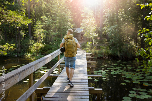 Mature man exploring Finnish nature in summer, walking across the bridge. Hiker with big backpack traveling in forests. Summer Scandinavian landscape of lakes and woods. 