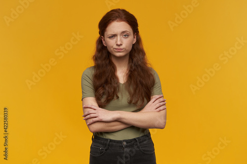 Unhappy looking girl, distrustful redhead woman with long hair. Wearing green t-shirt. People and emotion concept. Holding arms crossed. Watching at the camera, isolated over orange background