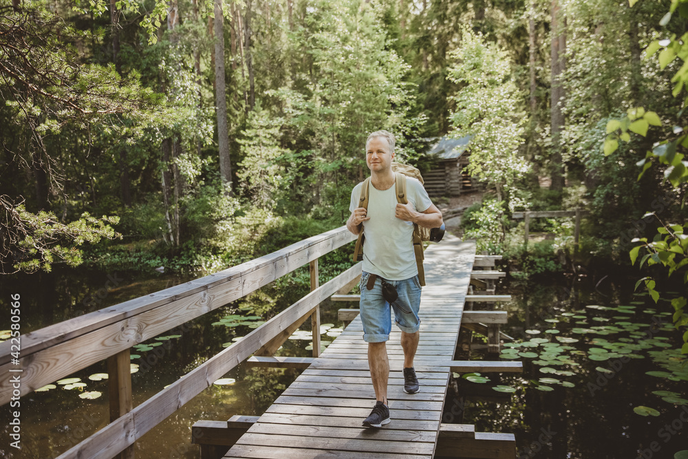 Mature man exploring Finnish nature in summer, walking across the bridge. Hiker with big backpack traveling in forests. Summer Scandinavian landscape of lakes and woods. 