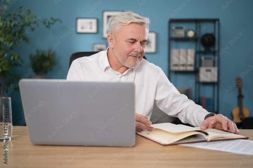 Older man working online with laptop computer at home sitting at desk. Home office, browsing internet, study room. Portrait of mature age, middle age, mid adult man in 50s. Older man working online with laptop computer at home sitting at desk. Home office, browsing internet, study room. Portrait of mature age, middle age, mid adult man in 50s.