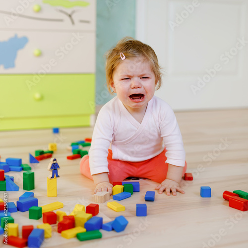 Upset crying baby girl with educational toys. Sad tired or hungry alone healthy child sitting near colorful different wooden blocks at home or nursery. Baby missing mother in daycare