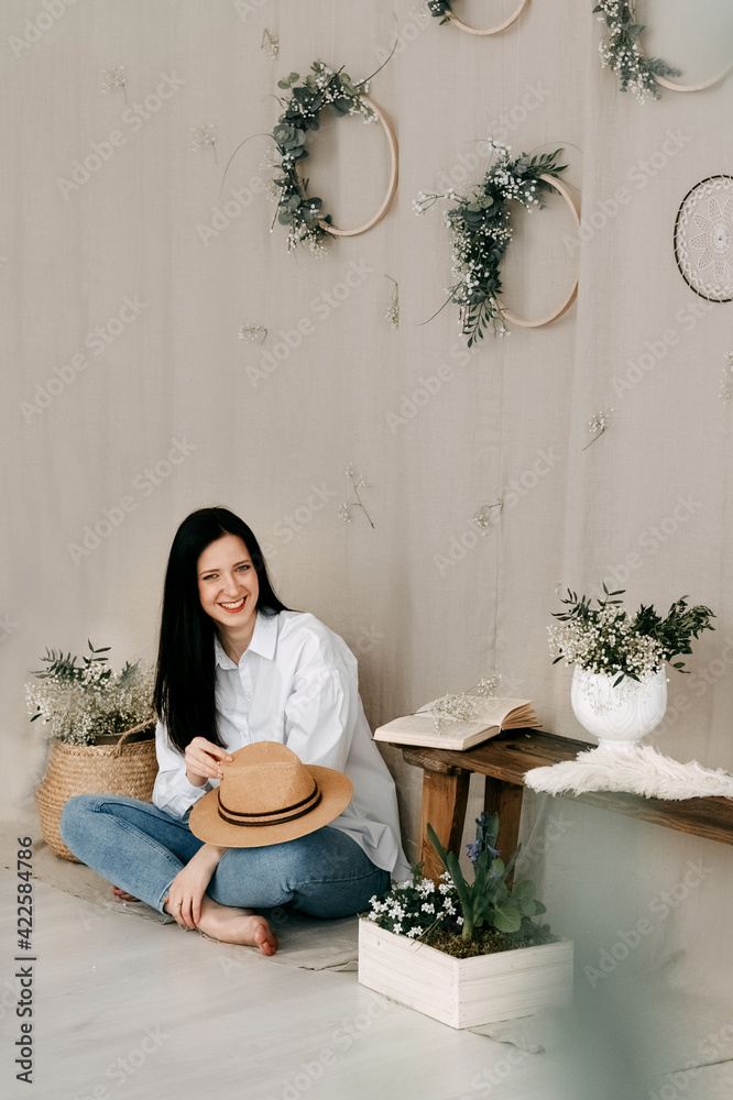 Laughing long-haired girl sits barefoot on the floor holding a hat in her hand. Behind her is a linen background with compositions of spring flowers. Laughing long-haired girl sits barefoot on the floor holding a hat in her hand. Behind her is a linen background with compositions of spring flowers.