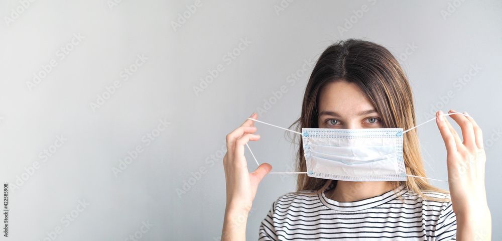 Young woman putting on medical surgical protection mask on grey background. Girl in hygienic face mask to prevent infection COVID-19, virus, coronavirus,allergy. Quarantine. Banner place for text Young woman putting on medical surgical protection mask on grey background. Girl in hygienic face mask to prevent infection COVID-19, virus, coronavirus,allergy. Quarantine. Banner place for text