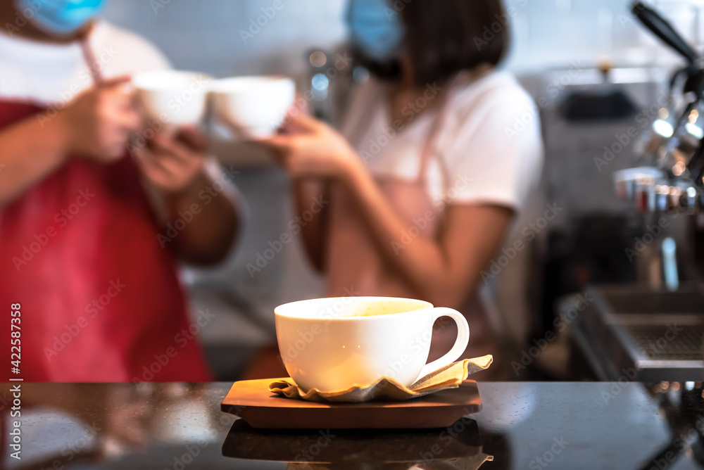 A white coffee cup Put on the counter with blur of two coffee barista wearing a surgical mask background, to people and coffee drink concept.