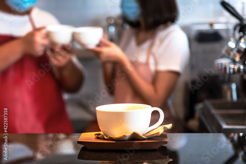 A white coffee cup Put on the counter with blur of two coffee barista wearing a surgical mask background, to people and coffee drink concept.