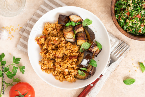 Bulgur pilaf (pilavi) with tomatoes and fried eggplants in a white plate on a light table top view. Turkish cuisine, vegan food.