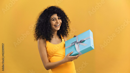 Happy pregnant hispanic woman holding gift box isolated on yellow