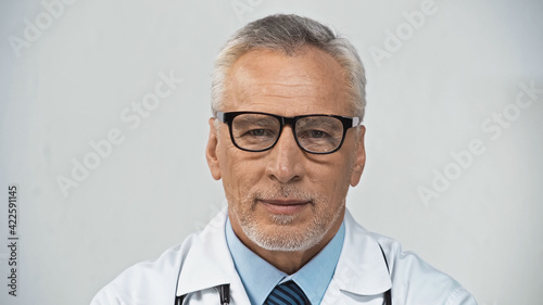 positive, mature doctor in eyeglasses smiling at camera in hospital