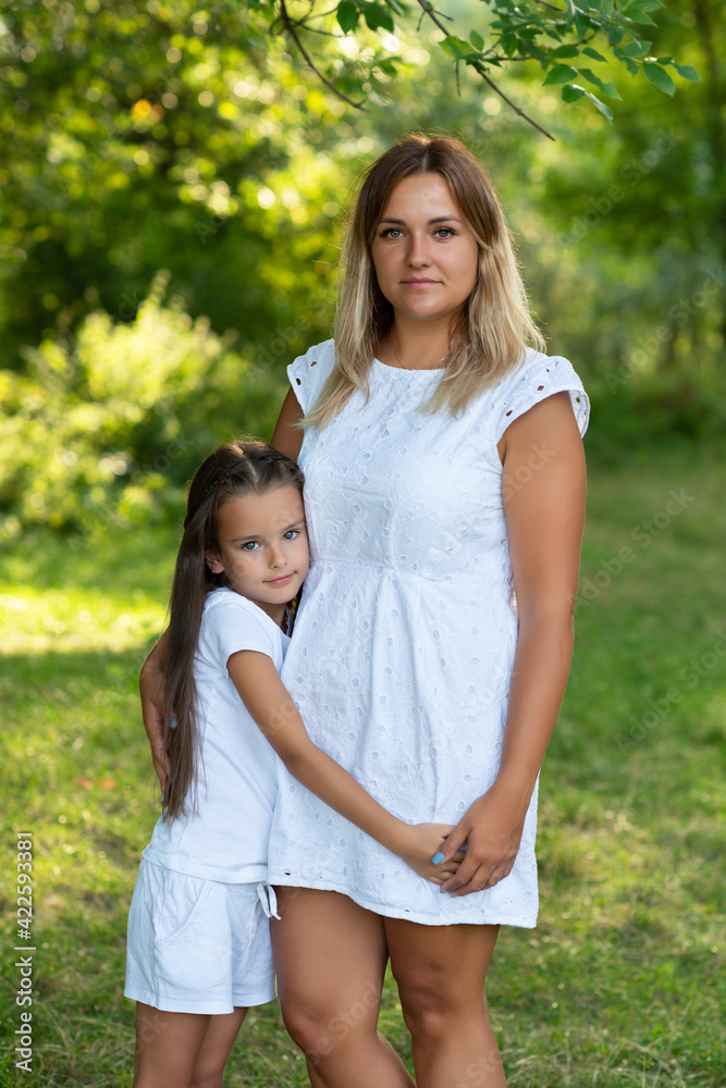 Obraz premium Little girl hugs her mother in summer forest nature outdoor. Portrait of mom and daughter wearing white clothes against summer greenery. 