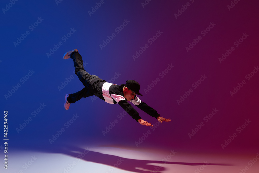 Beautiful sportive boy dancing hip-hop in stylish clothes on colorful gradient background at dance hall in neon light.