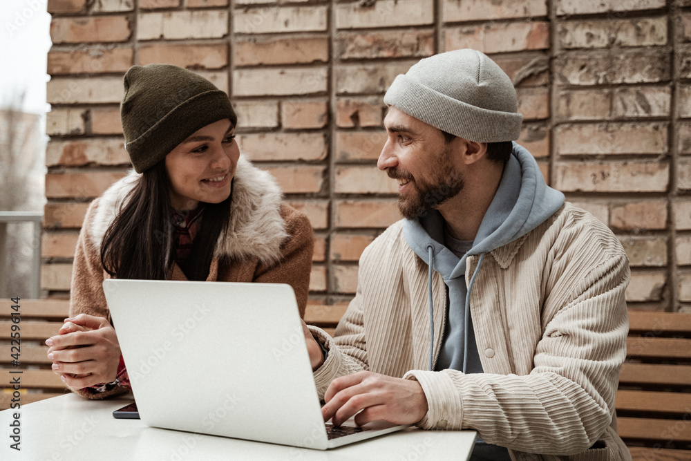 cheerful freelancer in beanie hats looking at each other near laptop cheerful freelancer in beanie hats looking at each other near laptop