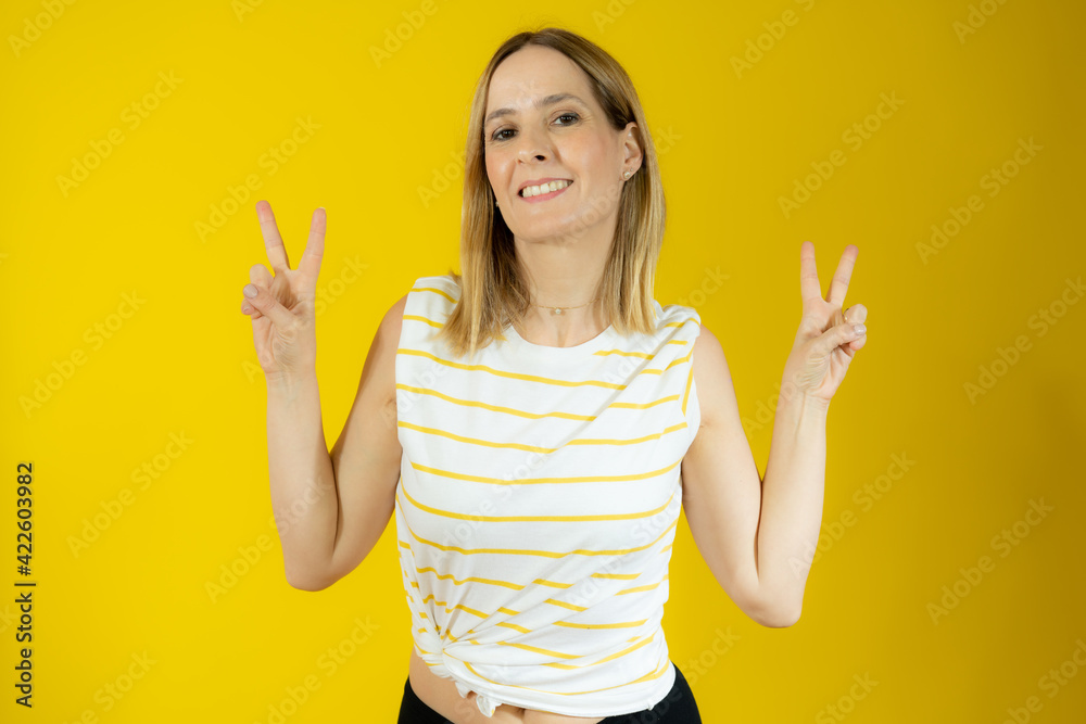 Portrait of a smiling woman showing victory sign and looking at camera isolated over yellow background