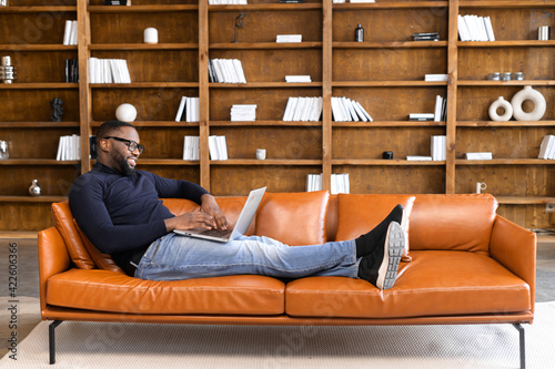 Cheerful African-American man wearing casual wear and eyeglases lying on the sofa and using laptop at modern home office, confident male entrepreneur typing on keyboard, a book shelves on background photo