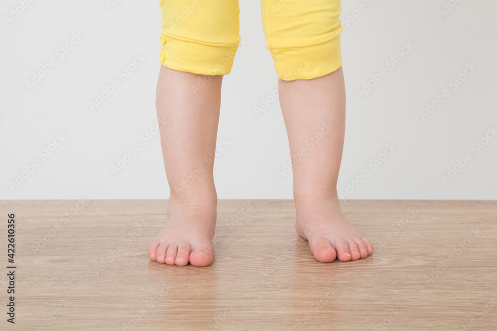 Baby girl barefoot standing on wooden floor at gray wall background. Closeup. Front view.