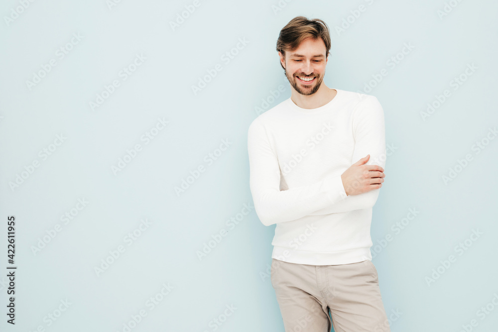 Portrait of handsome smiling hipster lumbersexual businessman model wearing casual white sweater and trousers. Fashion stylish man posing against light blue wall in studio Portrait of handsome smiling hipster lumbersexual businessman model wearing casual white sweater and trousers. Fashion stylish man posing against light blue wall in studio