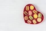 Multi colored french macaroons in a heart shape box top view on white wooden background.