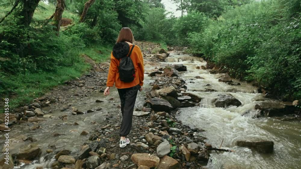 Woman tourist in an orange sweatshirt carefully walks on the stone of a mountain stream and looks at her legs, carries a backpack and a panama. Hiking in the mountains concept Woman tourist in an orange sweatshirt carefully walks on the stone of a mountain stream and looks at her legs, carries a backpack and a panama. Hiking in the mountains concept
