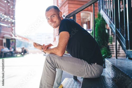 Portrait of cheerful Caucasian male blogger with touch pad smiling at camera enjoying time for networking social media, joyful hipster guy with digital tablet or ebook posing at urban setting