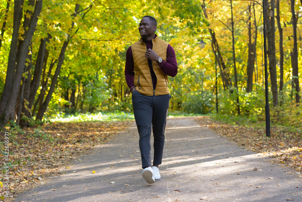 Positive african american young man in stylish clothes walks in late summer park on sunny warm warm day. Outdoor activity concept. Positive african american young man in stylish clothes walks in late summer park on sunny warm warm day. Outdoor activity concept.