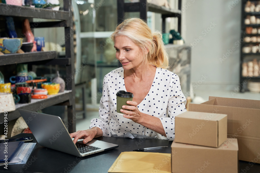 Busy mature woman, small business owner smiling and drinking coffee while working on the laptop, sitting at the desk in her craft pottery shop Busy mature woman, small business owner smiling and drinking coffee while working on the laptop, sitting at the desk in her craft pottery shop