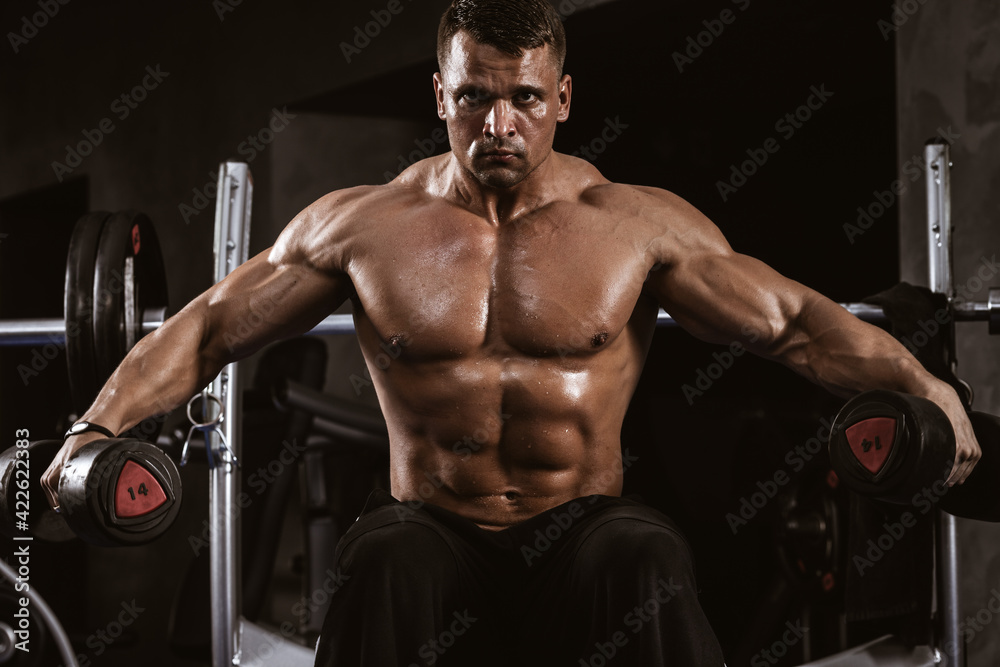 Fitness in gym, sport and healthy lifestyle concept. Handsome athletic man with naked torso making exercises. Bodybuilder male model trains deltoid muscles with dumbbells. Heavy barbell on background