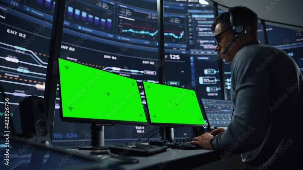 Professional IT Technical Support Specialist and Software Developer Working on Computer with Green Screen Mock Up Display in Monitoring Control Room with Digital Screens. Employee Wears Headphones.
