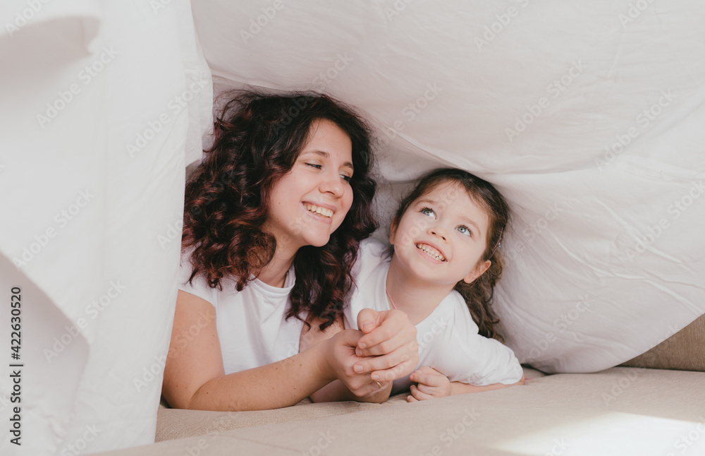 a happy little girl in a light turtleneck hugs her mother under a white sheet. a loving and caring mother a happy little girl in a light turtleneck hugs her mother under a white sheet. a loving and caring mother