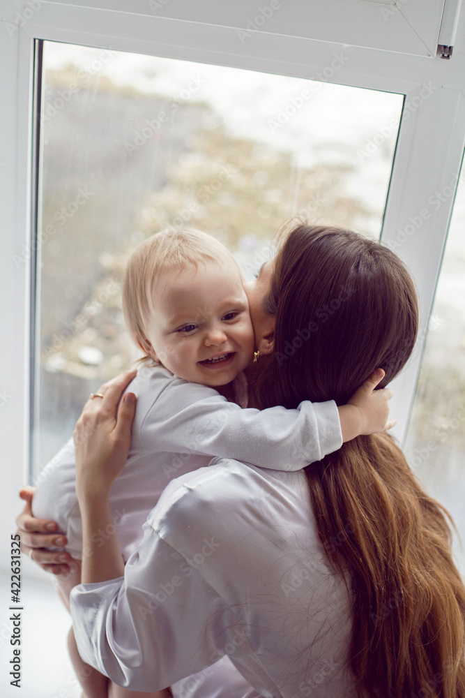 mother in a white robe sits with a child a blonde daughter at a large window of the house