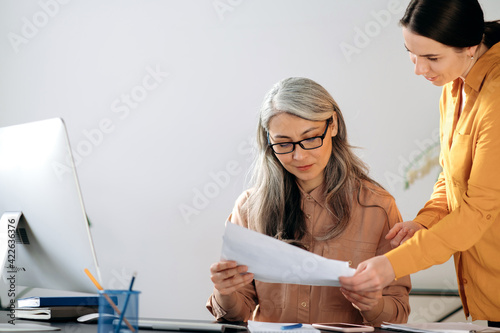 Friendly attractive confident mature Asian woman wearing glasses, ceo, manager, business woman working in modern office wearing stylish clothes, looks at female employee financial statement