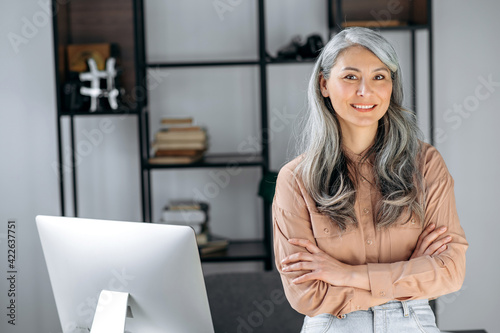 Portrait of a beautiful successful confident gray-haired Asian woman, business lady, top manager, stands in a modern office with arms crossed, in stylish clothes, looks at the camera, smiling friendly