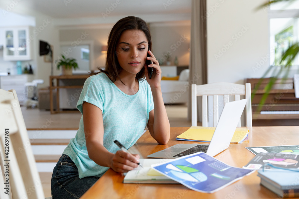 Caucasian woman sitting by desk working from home talking on smartphone and using laptop Caucasian woman sitting by desk working from home talking on smartphone and using laptop