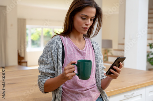 Caucasian woman standing in kitchen drinking cup of coffee using smartphone and smiling