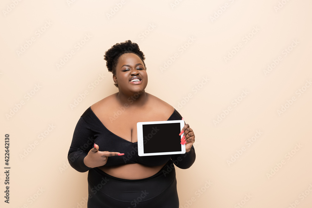 happy african american plus size woman pointing with finger at digital tablet with blank screen isolated on beige happy african american plus size woman pointing with finger at digital tablet with blank screen isolated on beige
