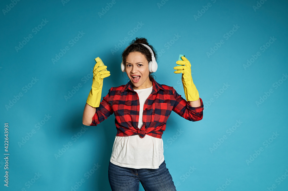 Young african woman in red plaid shirt with headphones on her head sings, posing on a blue background with cleaning sponges in her hands