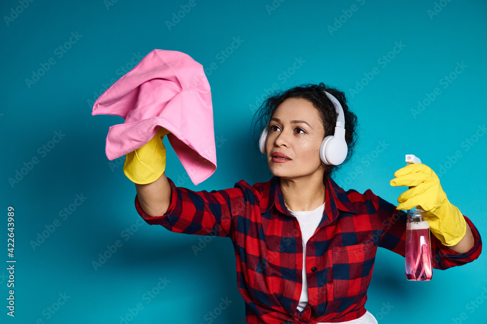 Woman in headphones and yellow rubber gloves holds detergent spray and cleaning rag, simulating cleaning surface. Woman in headphones and yellow rubber gloves holds detergent spray and cleaning rag, simulating cleaning surface.