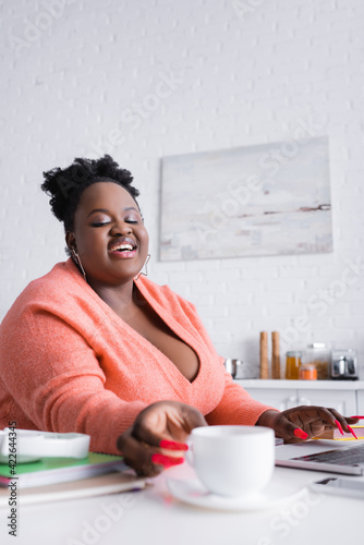 happy african american plus size woman reaching cup of coffee on blurred foreground
