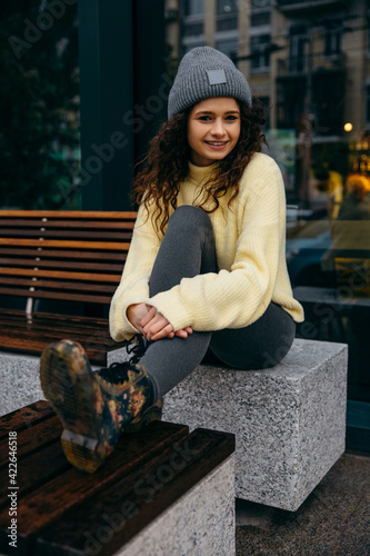 Pretty young curly girl in yellow sweater and gray cap sitting at the city bench. photo