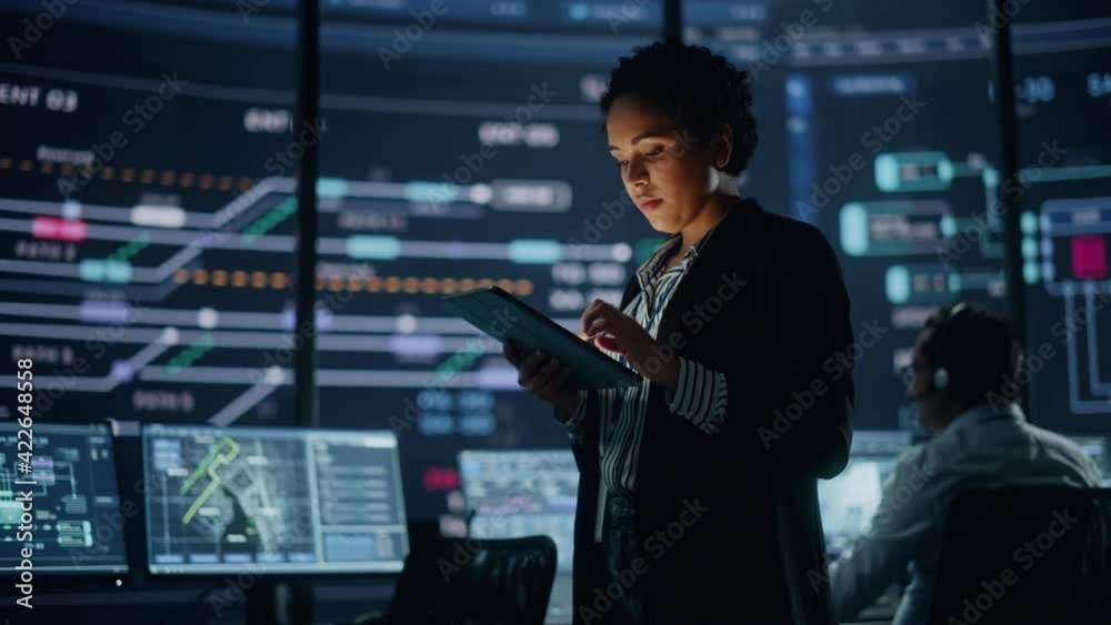 Young Multiethnic Female Government Employee Uses Tablet Computer in System Control Monitoring Center. In the Background Her Coworkers at Their Workspaces with Many Displays Showing Technical Data.