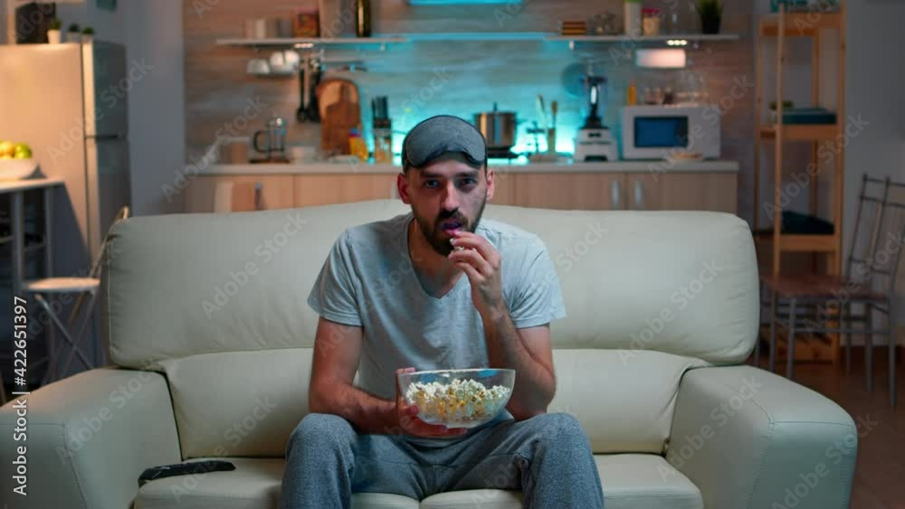 Concentrated man with sleep eye mask sitting in front of television watching lifestyle show. Caucasian male relaxing on couch holding popcorn bowl late at night in kitchen Concentrated man with sleep eye mask sitting in front of television watching lifestyle show. Caucasian male relaxing on couch holding popcorn bowl late at night in kitchen