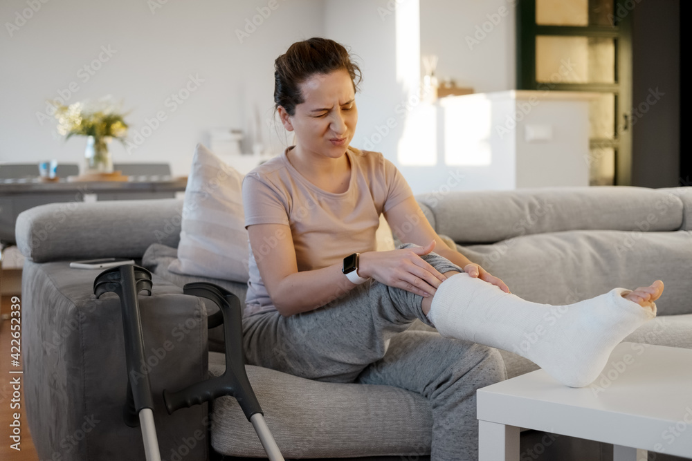 Obraz premium Young woman with pain expression on couch at home with orthopedic plaster massaging leg. Fracture of the leg or foot. Concept of rehabilitation and healing physiotherapy. Orthopedics and Traumatology.