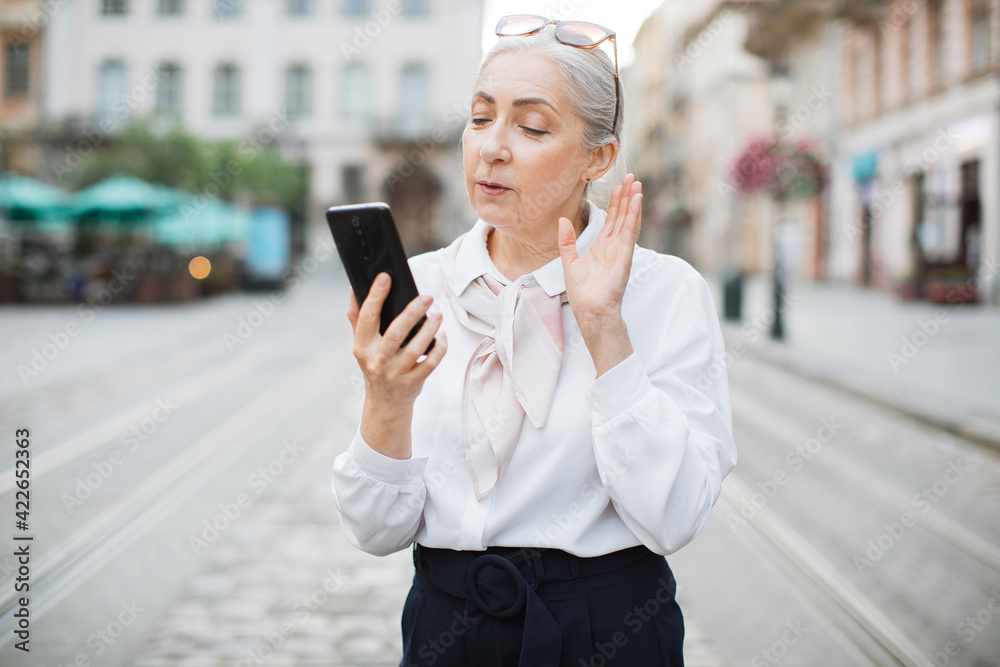 Happy mature lady in stylish outfit talking and waving during video conversation on smartphone. Positive senior woman standing on city street and using modern gadget. Happy mature lady in stylish outfit talking and waving during video conversation on smartphone. Positive senior woman standing on city street and using modern gadget.