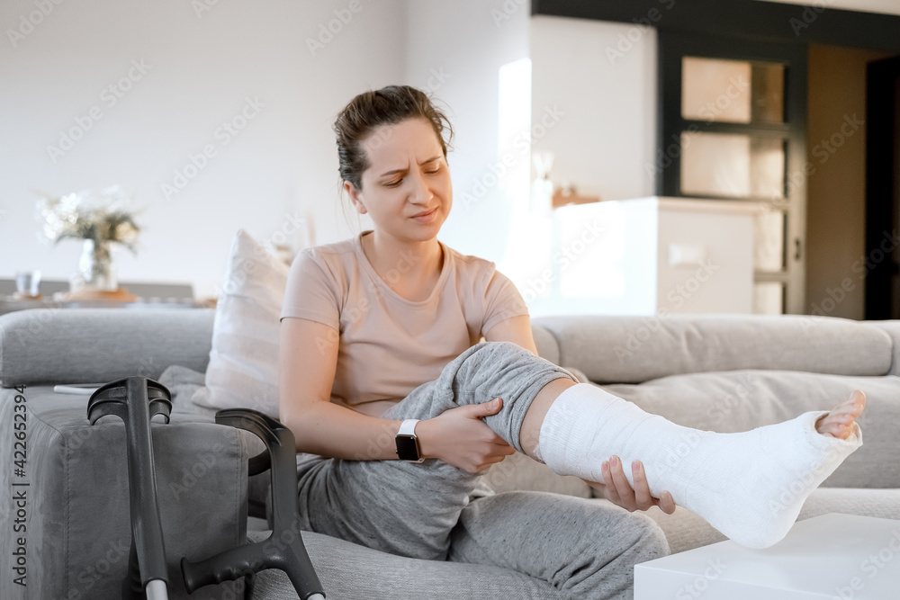 Young woman with pain expression on couch at home with orthopedic plaster massaging leg. Fracture of the leg or foot. Concept of rehabilitation and healing physiotherapy. Orthopedics and Traumatology. Young woman with pain expression on couch at home with orthopedic plaster massaging leg. Fracture of the leg or foot. Concept of rehabilitation and healing physiotherapy. Orthopedics and Traumatology.