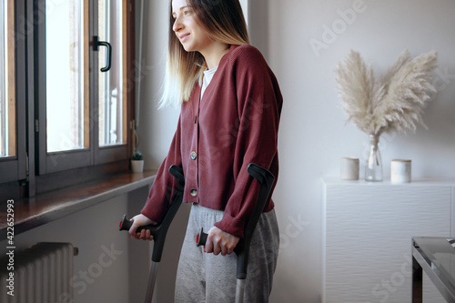 Obraz na płótnie Adult woman in her late twenties on crutches at home is looking into the window with hope