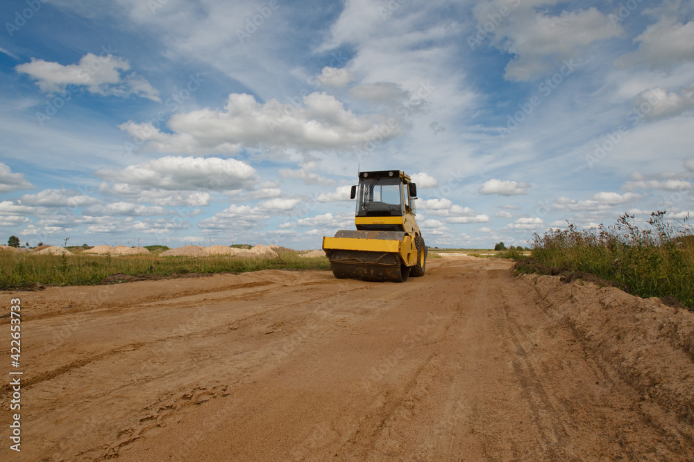 Single drum roller during work. He is moving along ground road. Concept - laying a country road. Road construction. Single drum roller services. Driveway paving works on a summer day.