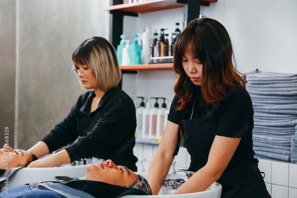 Fototapeta premium Serious Asian female hairdresser wearing uniform and spectacles shampooing hair of young woman customer in modern salon using organic shampoo and conditioner