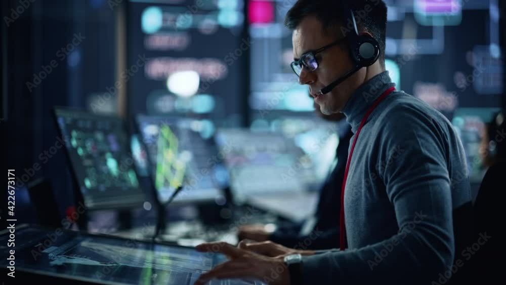 Portrait of Professional IT Technical Support Specialist Working on Computer in Monitoring Control Room with Digital Screens. Employee Wears Headphones with Mic and Talking on a Call. Portrait of Professional IT Technical Support Specialist Working on Computer in Monitoring Control Room with Digital Screens. Employee Wears Headphones with Mic and Talking on a Call.