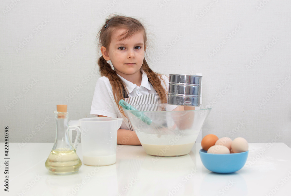 A little charming girl in an apron prepares dough from flour, eggs and milk, child mixes ingredients in glass bowl, little cook prepares food. Homemade cakes