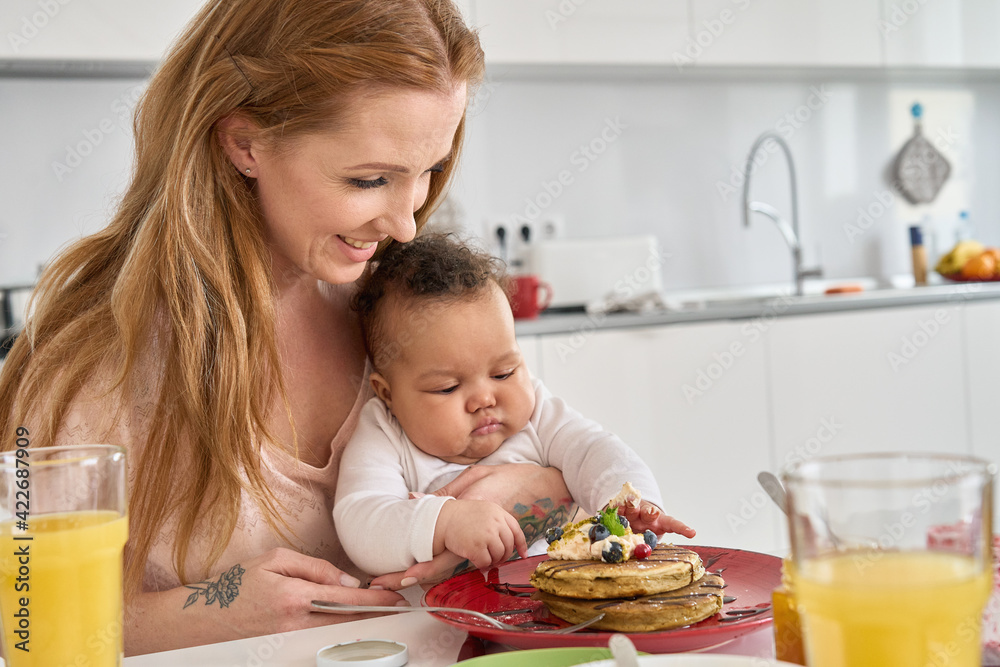 Cute mixed race baby daughter sitting on mum lap at kitchen table. Curious adorable infant child girl tasting pancakes while happy Caucasian mother playing with little kid having breakfast at home.