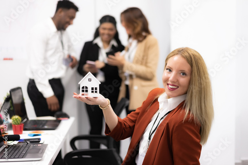 Real estate agent sustaining over hand a little white house, desktop with tools and laptops. Background there are business people working with smartphones. Technology, mortgage, rent and buy concept.